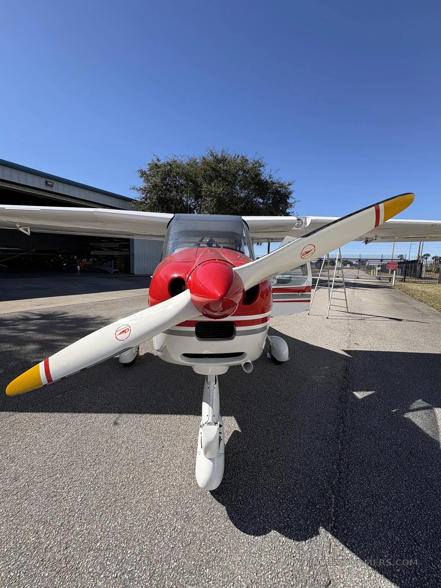A close-up, head-on view of a white and red Tecnam P2008 aircraft shows its propeller and nose cone, parked on an asphalt surface.