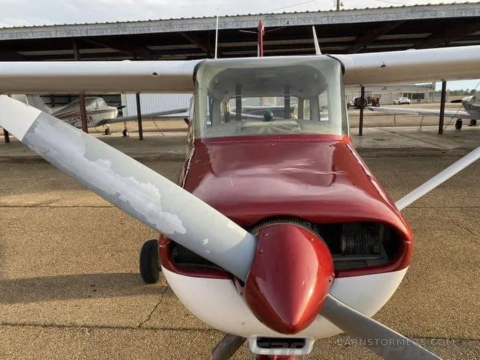 Front view of a red and white single-engine aircraft with a two-blade propeller, parked on an airfield.