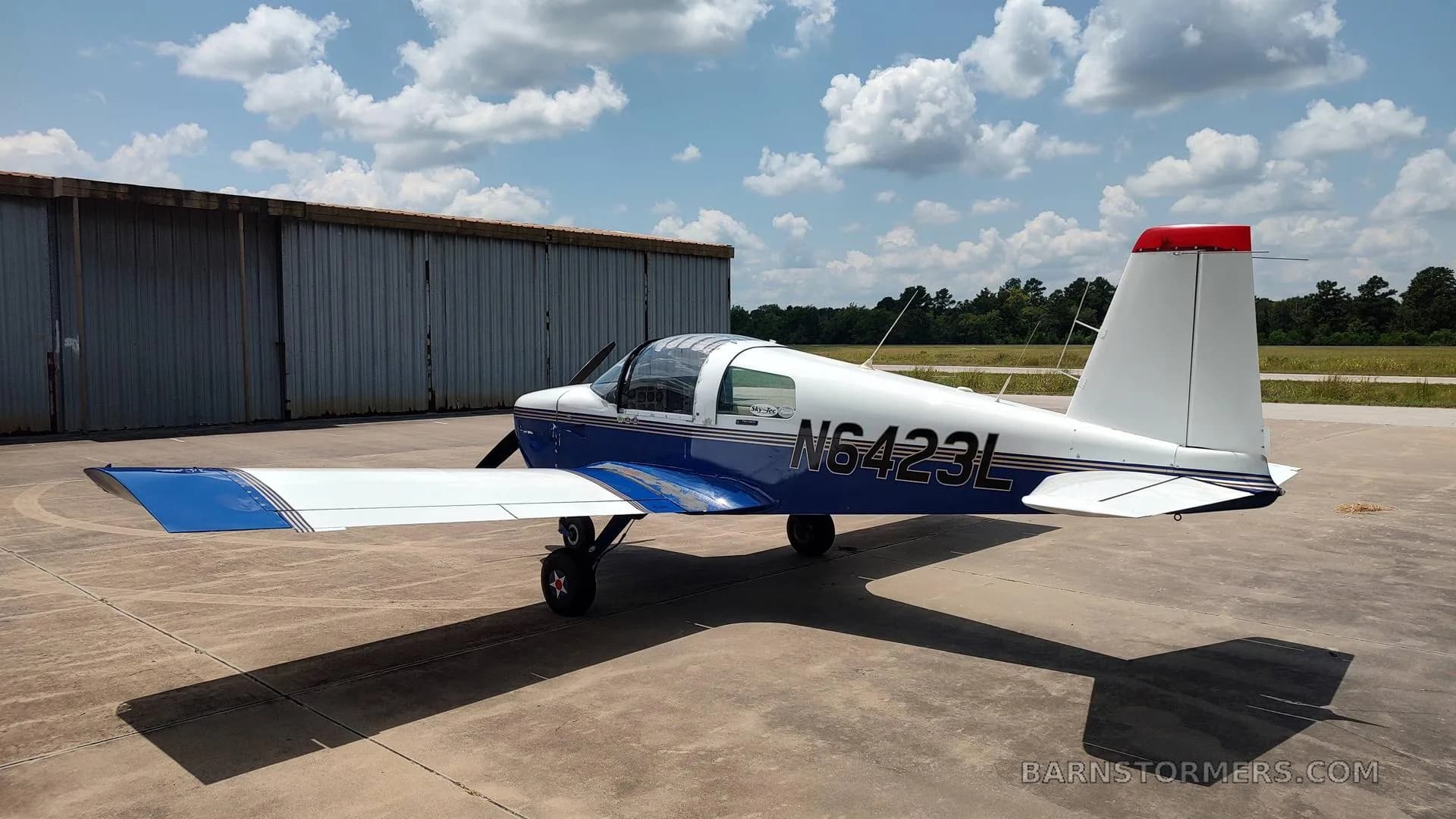 A white and blue American Aviation AA-1 Yankee aircraft with registration N6423L, viewed from the rear-right, parked on an asphalt surface next to a hangar.