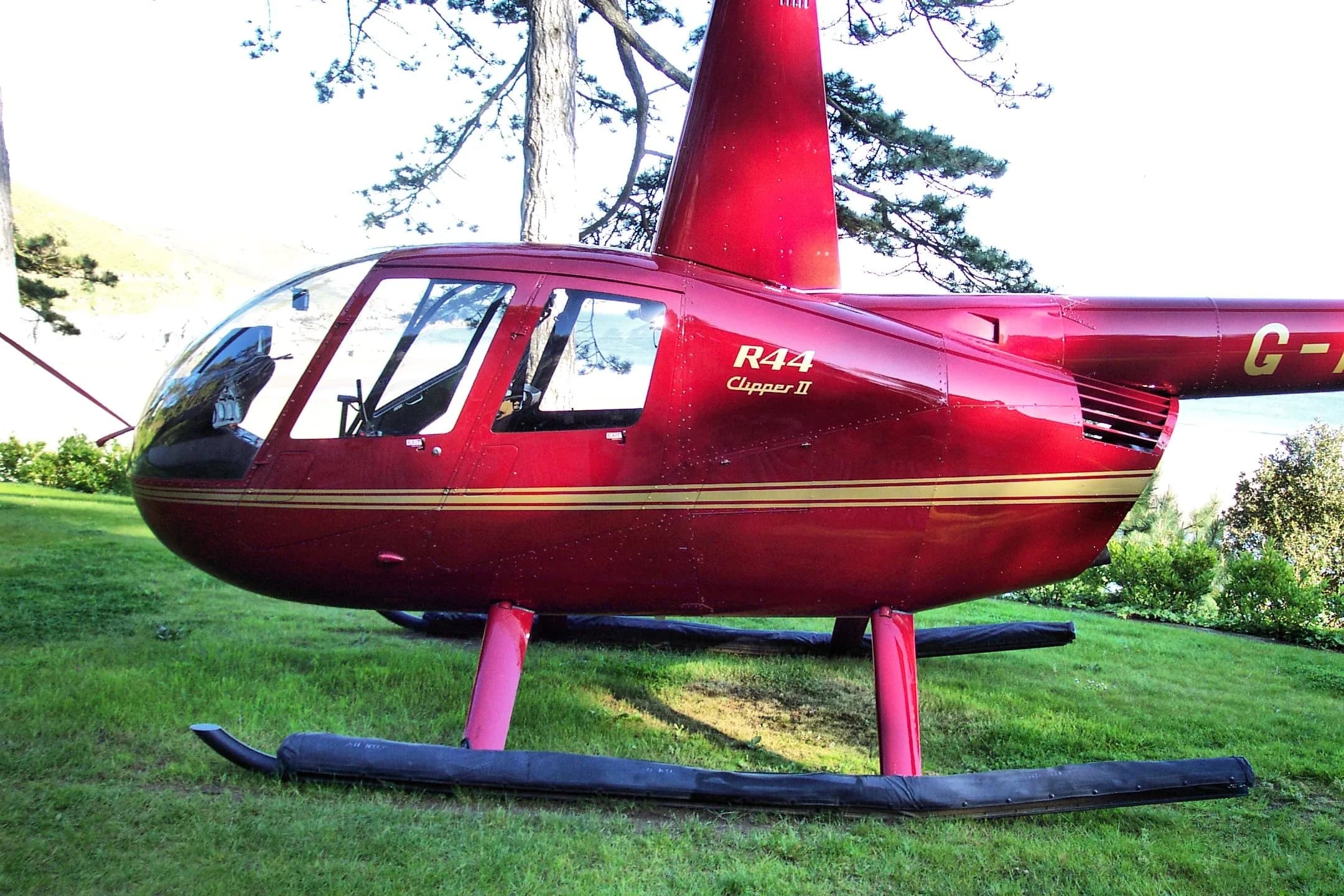 A glossy red Robinson R44 Clipper II helicopter with gold stripes, parked on a grassy lawn.