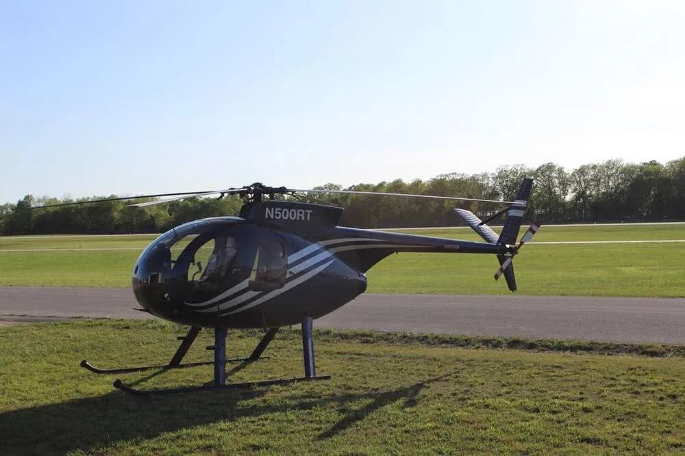 A blue and white Hughes 500 helicopter (N500RT) is parked on a grassy field next to a runway, viewed from the side.