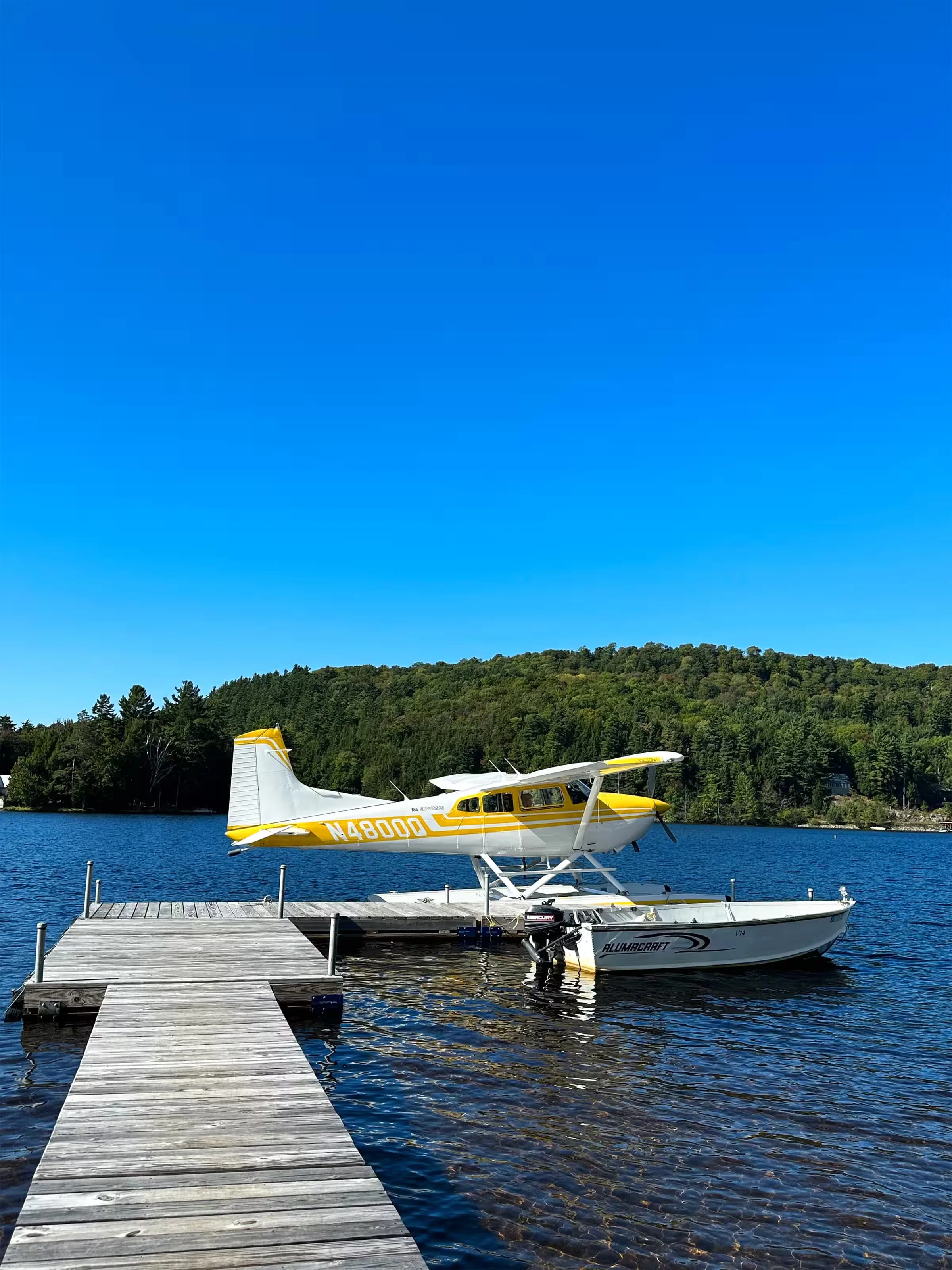 A yellow and white Cessna 185 floatplane, N4800Q, docked at a wooden pier on a clear lake under a blue sky.
