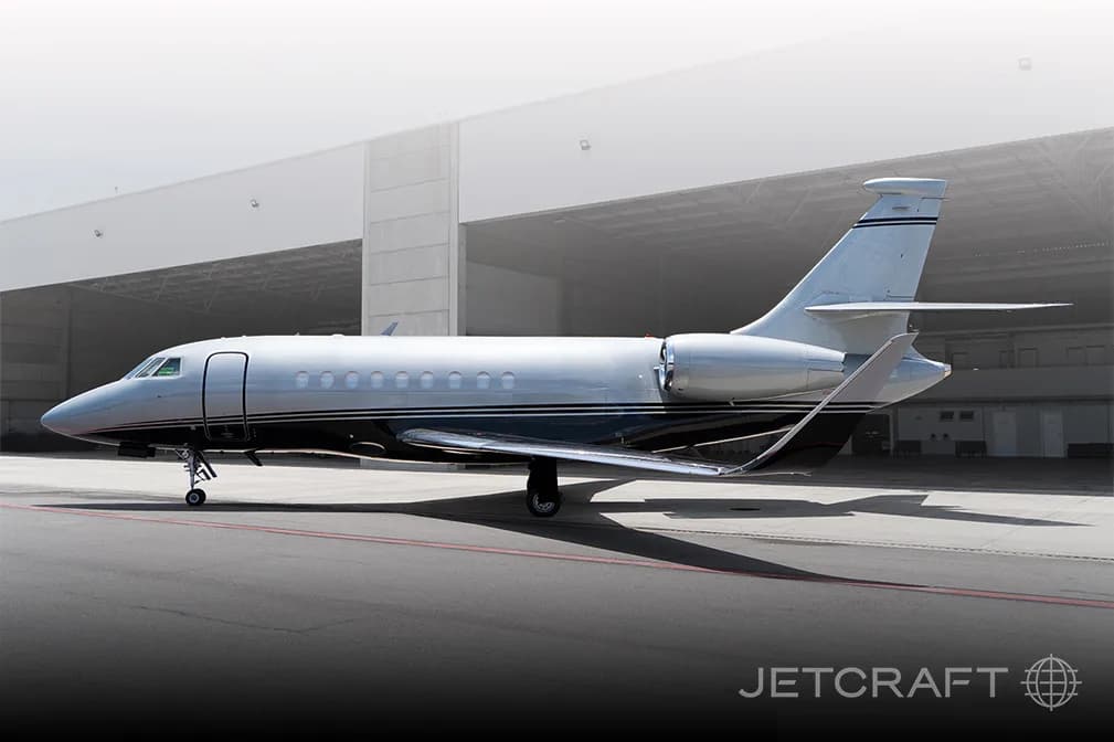 A sleek silver and black business jet is parked on an asphalt tarmac next to a large hangar, viewed from the side.