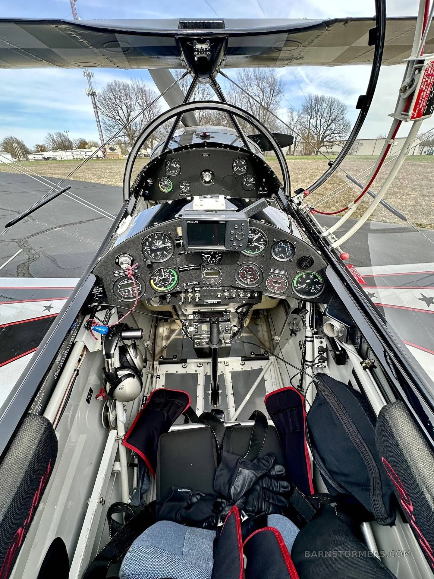 The cockpit of a Pitts biplane, showing the instrument panel with gauges and a central display, and two seats with harnesses.