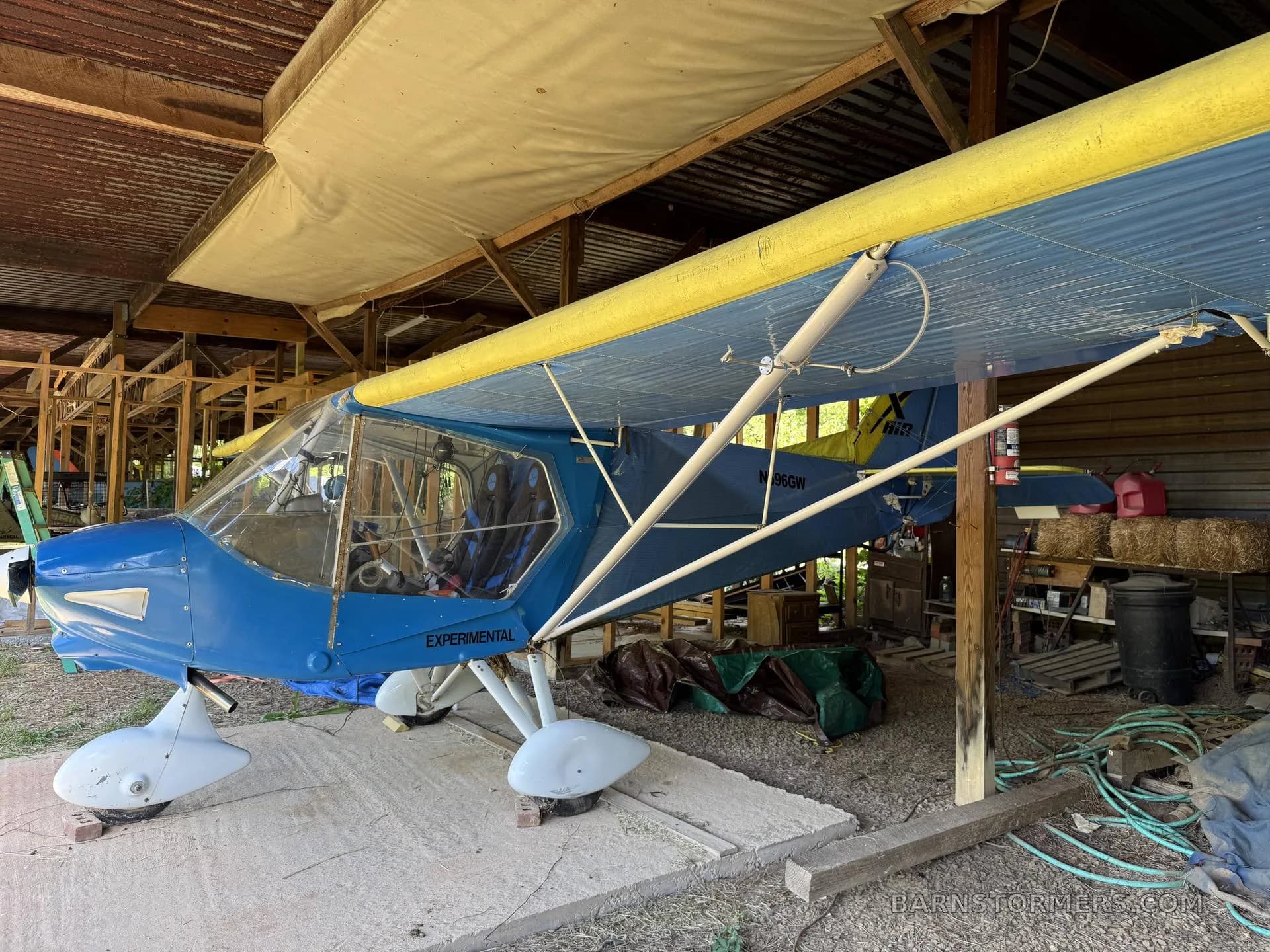 A blue and yellow experimental aircraft with white floats is parked inside a rustic shed, viewed from the side.