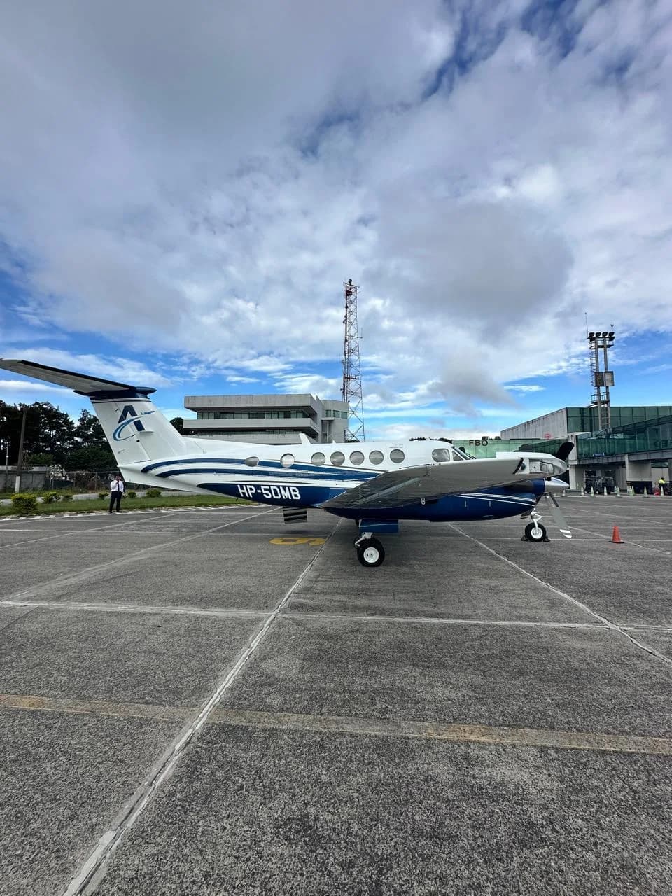 A white and blue turboprop aircraft (King Air) with registration HP-5DMB parked on an airport tarmac with airport buildings and communication towers in the background.