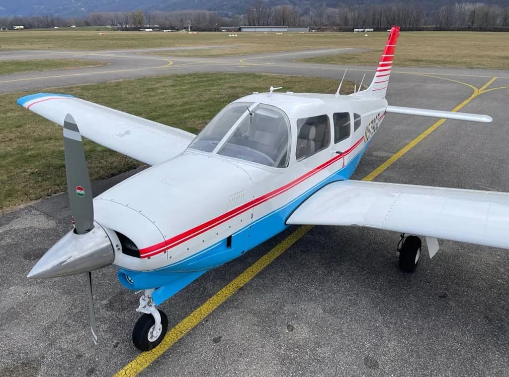 A front-right view of a white and blue Piper PA-28-181 Archer II with red stripes, parked on an airfield.
