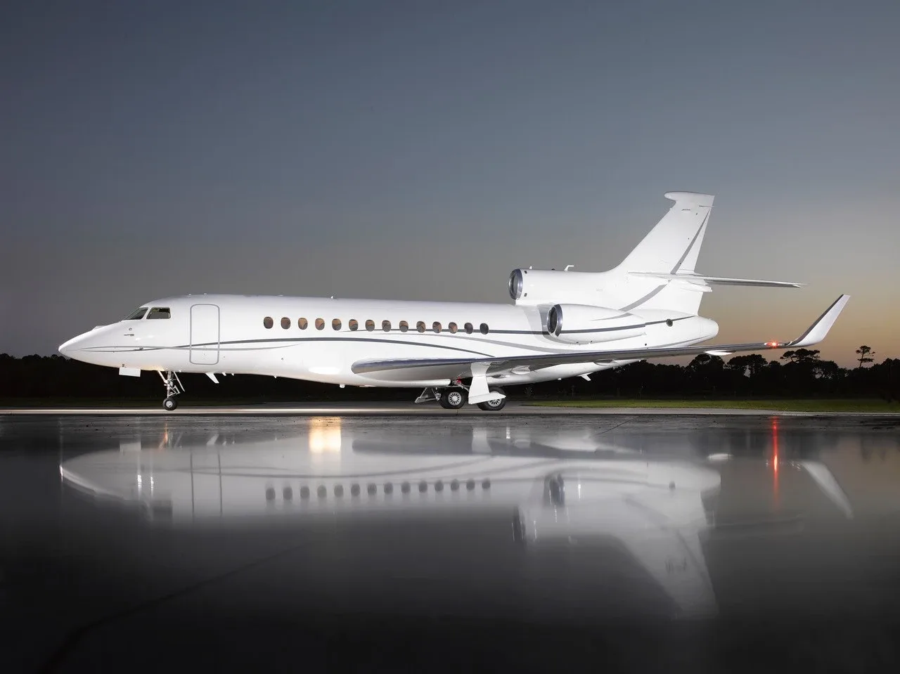 A white Dassault Falcon 7X business jet, registration N7X, parked on a wet tarmac at dusk, reflecting in the water.