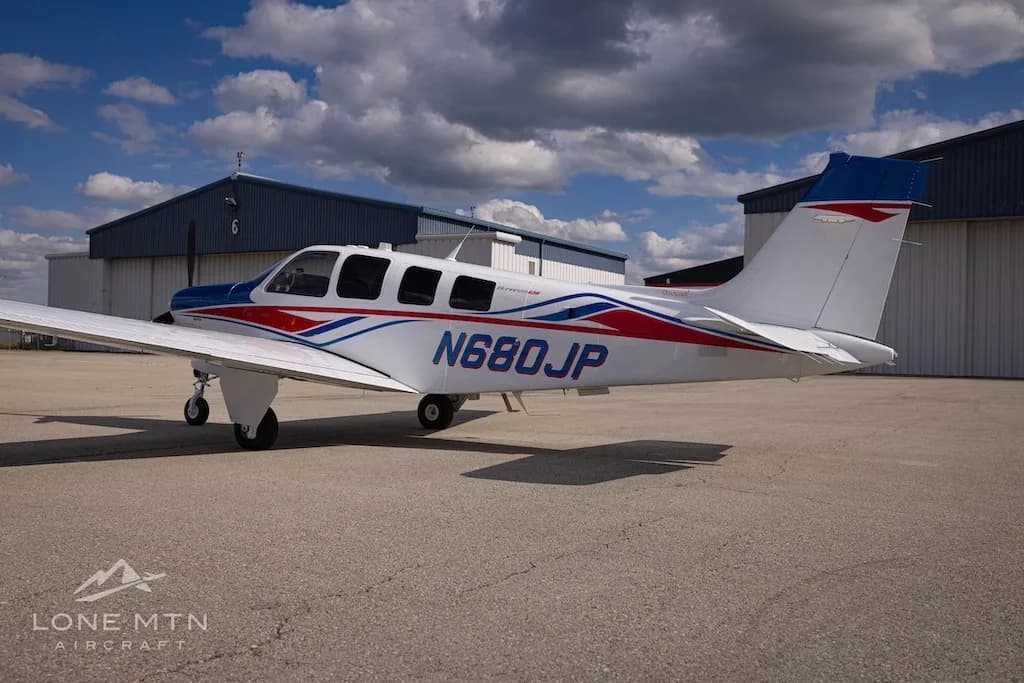 A side view of a white, blue, and red Beechcraft Bonanza N680JP parked on an airfield next to a hangar.