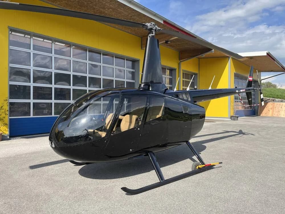 A black Robinson R44 helicopter with tan interior seats is parked on an asphalt apron in front of a yellow hangar.