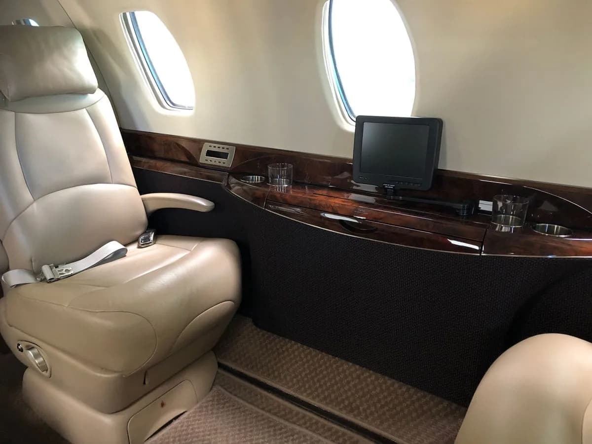 A close-up of a beige leather seat in a private jet cabin with a window and a small entertainment screen.
