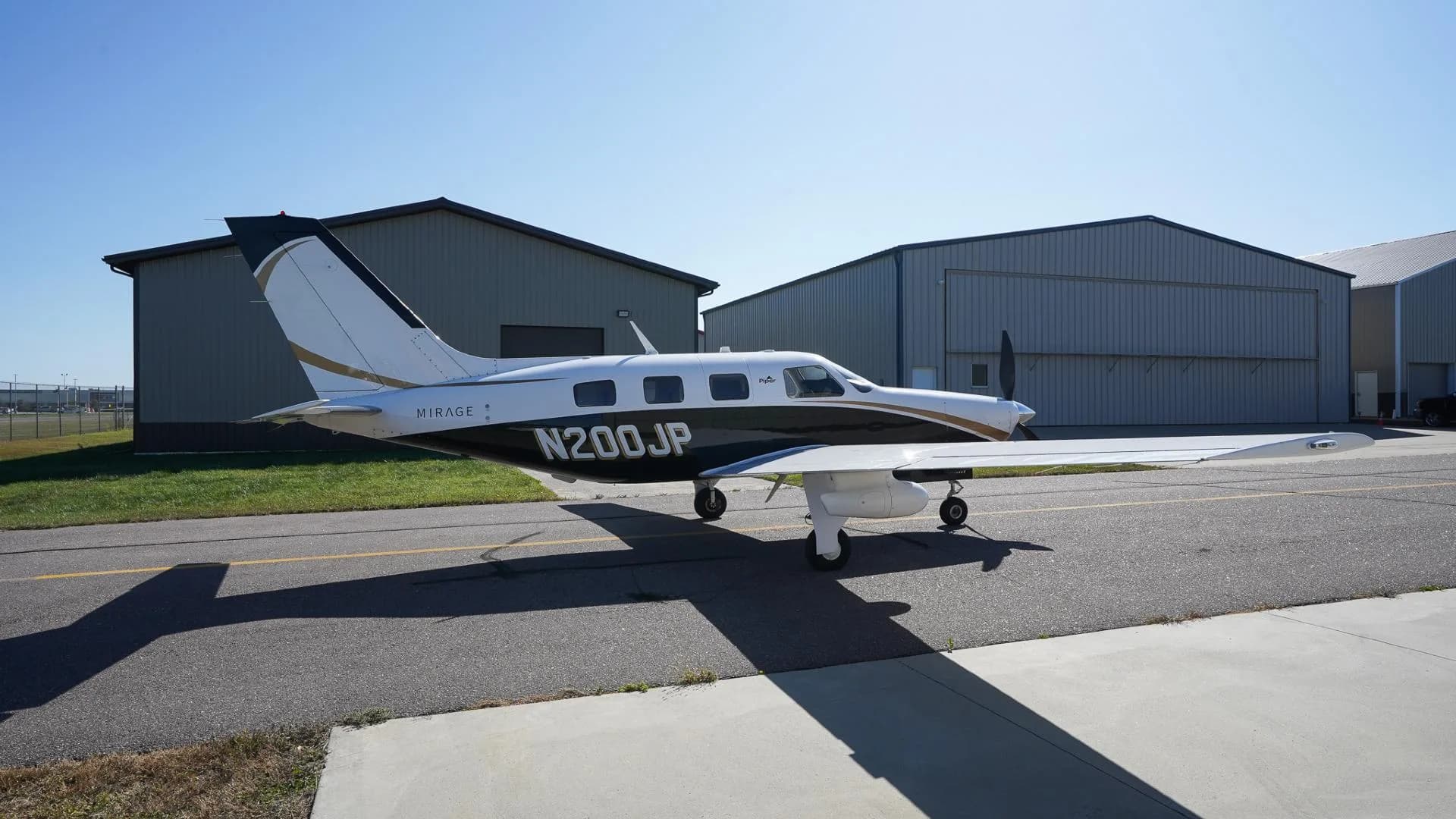 A white and black Piper Mirage N200JP with gold accents parked on an airfield next to hangars.