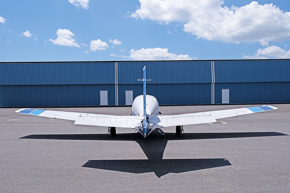 Rear view of a white and blue Mooney M20F aircraft parked on the tarmac in front of a hangar.