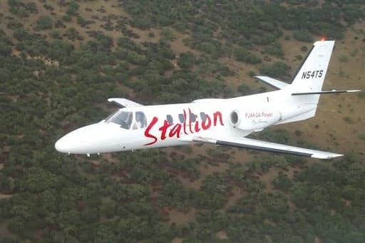 A white Stallion FJ44-2A private jet in flight over a wooded area.