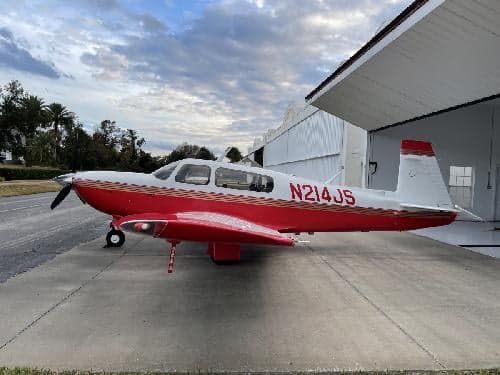 A red and white Mooney M20C aircraft, registration N214JS, parked outside a hangar.