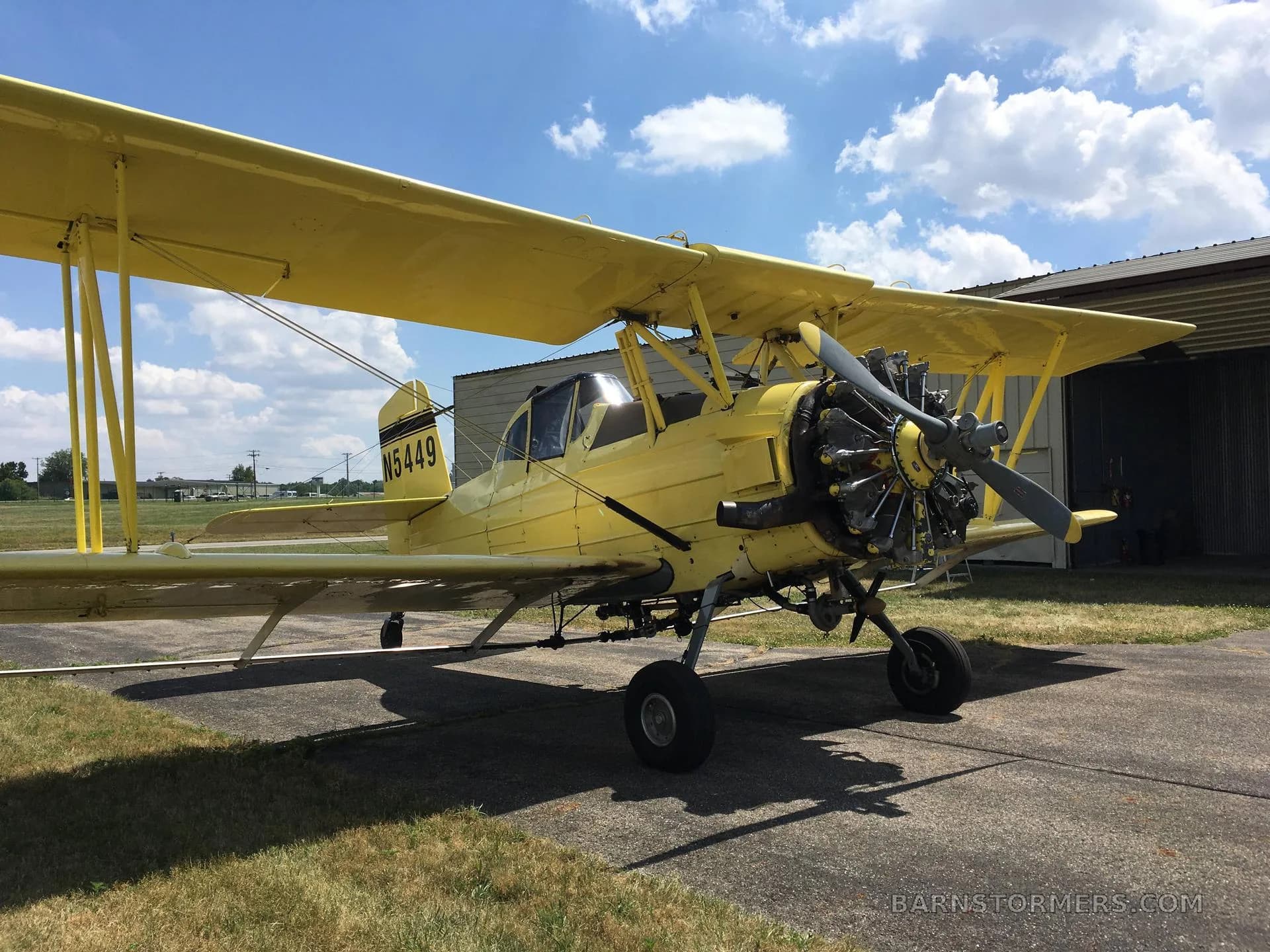 A yellow biplane crop duster with its radial engine visible, parked on an airfield with a hangar in the background.