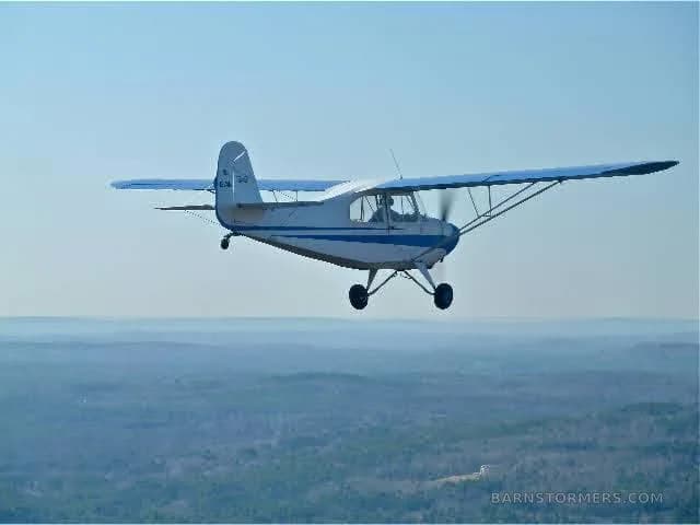 A white and blue single-engine high-wing aircraft, registration NC 1274E, flying over a green forested landscape under a blue sky.