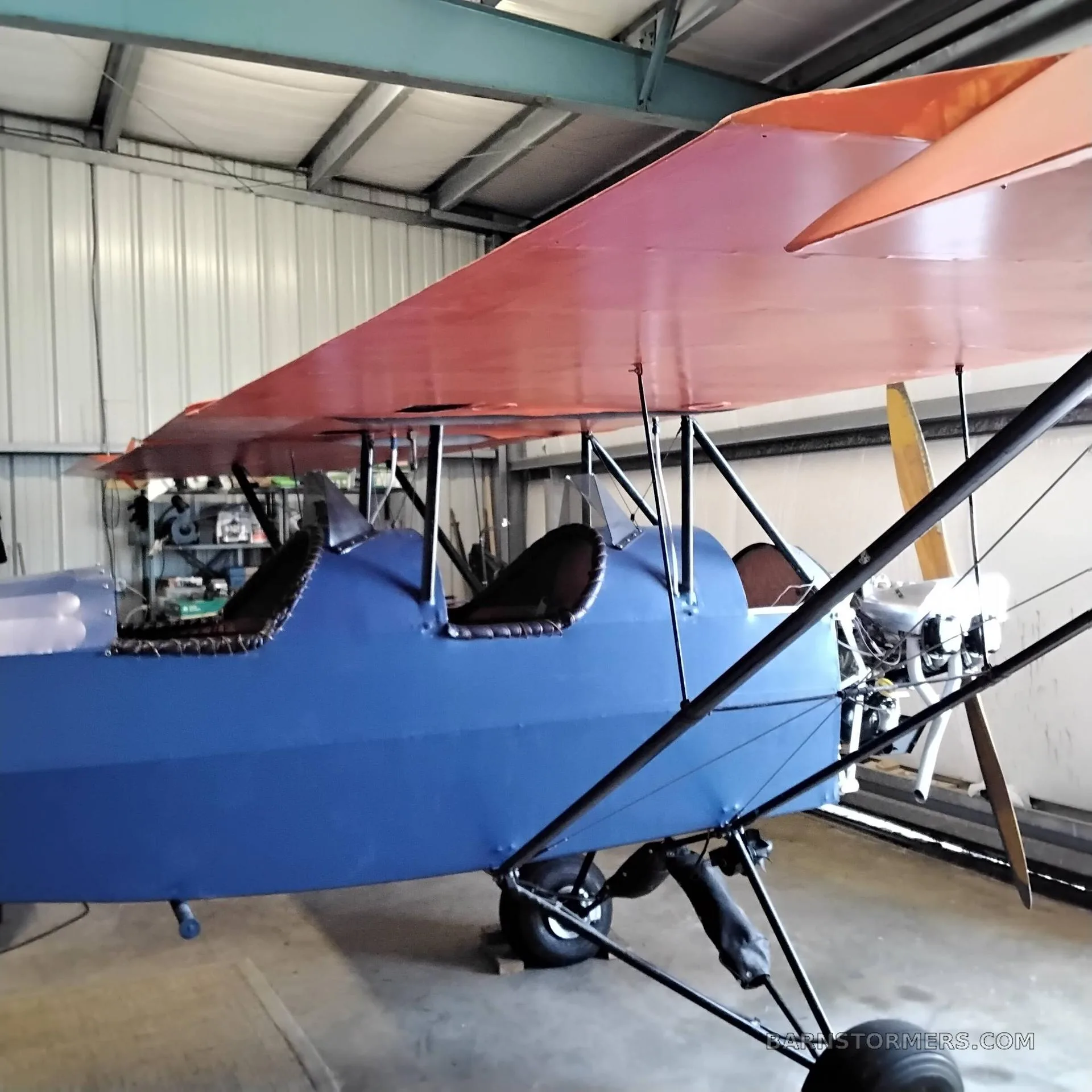 A side view of a blue and orange biplane with two open cockpits, parked inside a hangar.