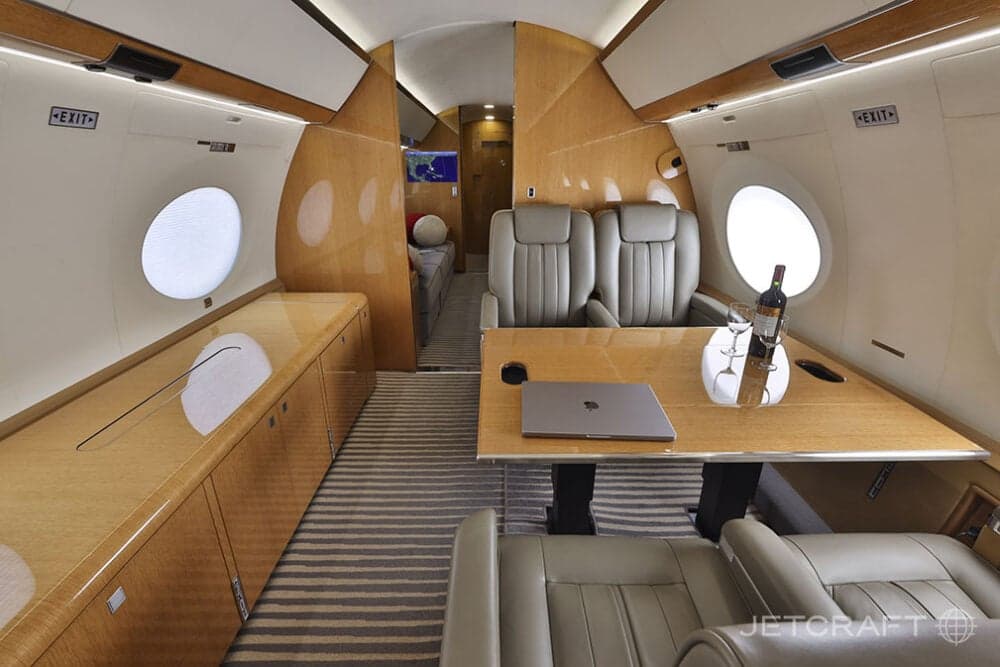 A wide view of the business jet cabin showing light brown leather seats, a wooden table with a laptop and wine, and a rear lounge area.