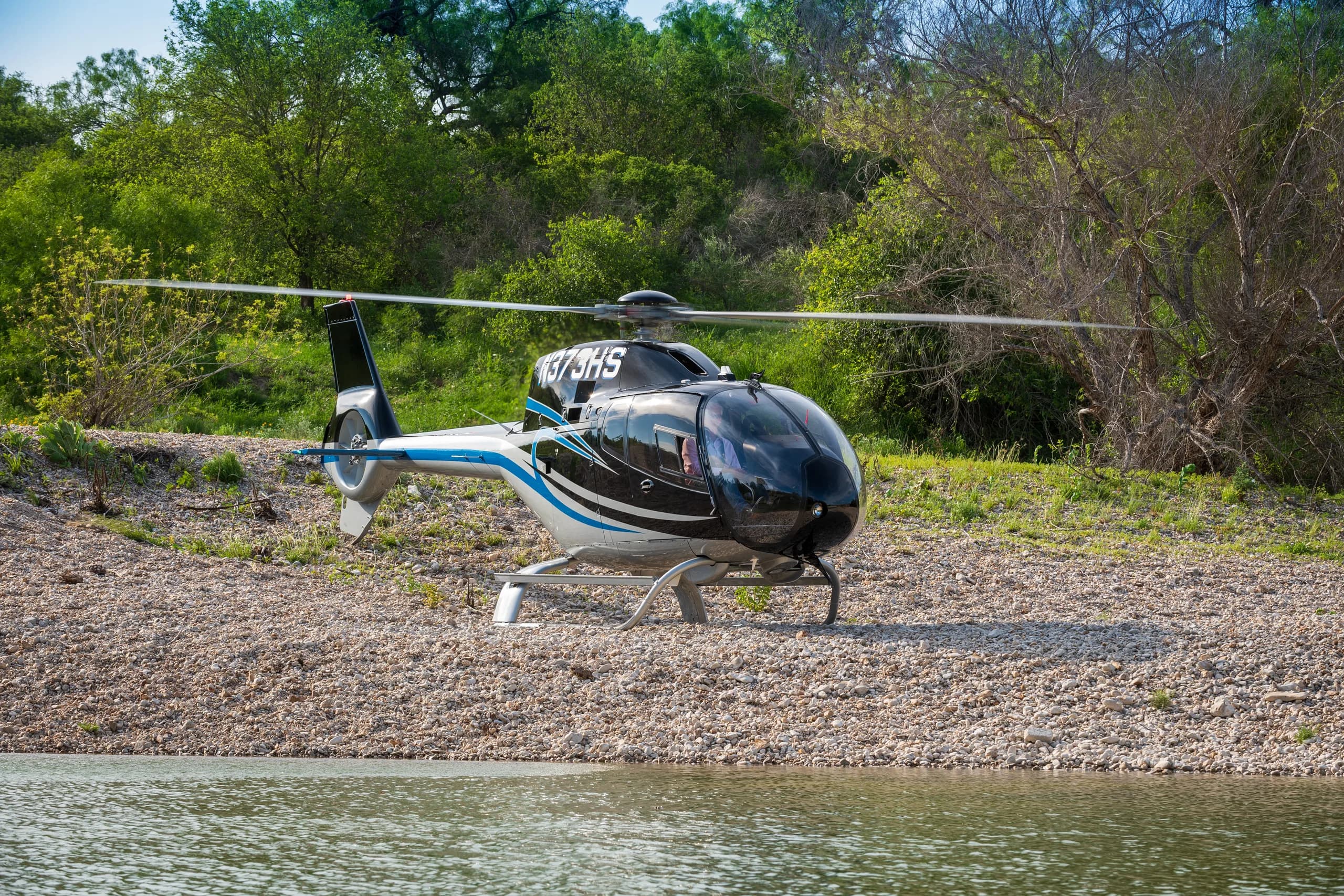 A black and blue helicopter (N373HS) with a white stripe sits on a rocky shore next to a body of water.