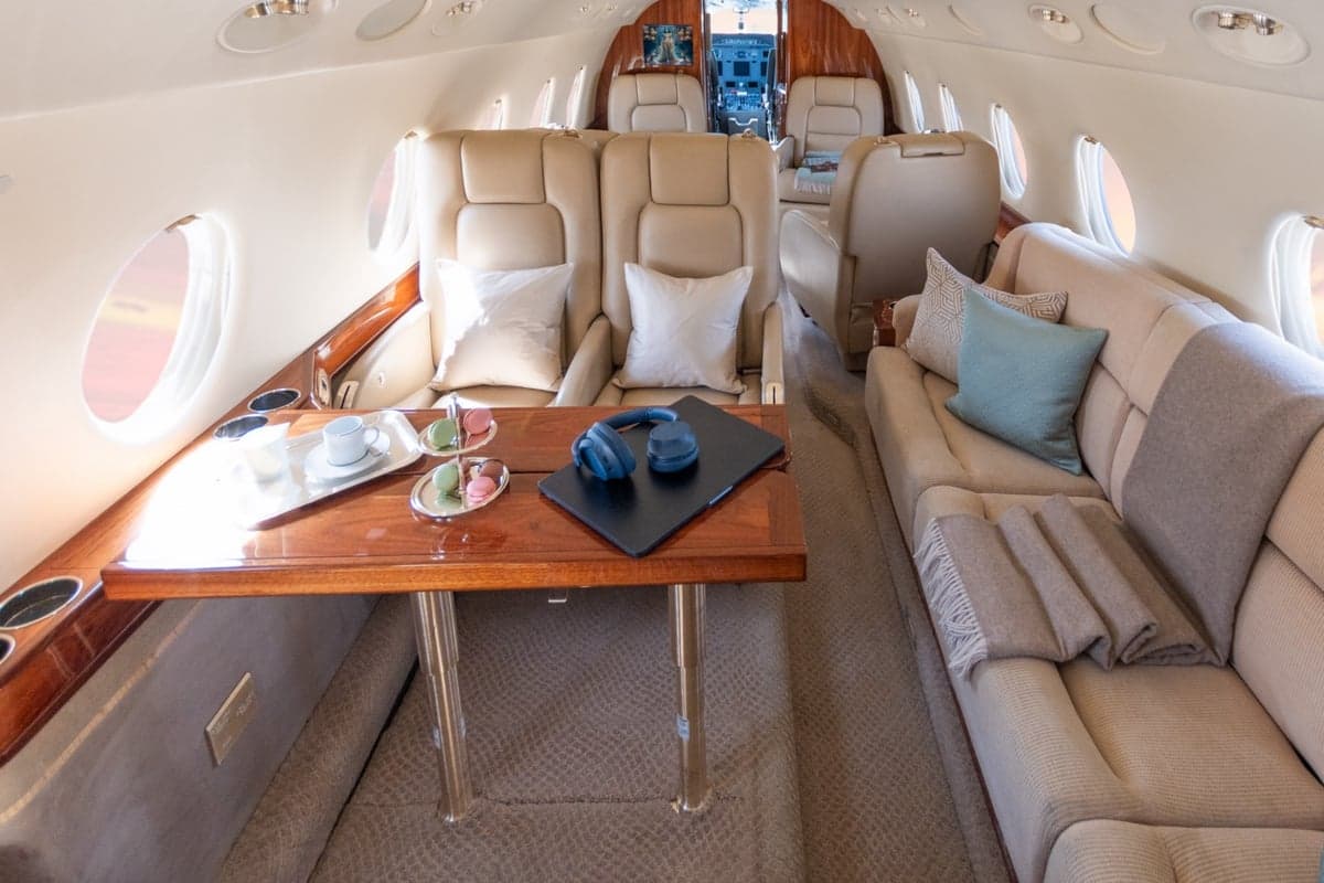 A spacious aircraft cabin interior featuring a long beige sofa, individual seats, and a wooden table with refreshments.
