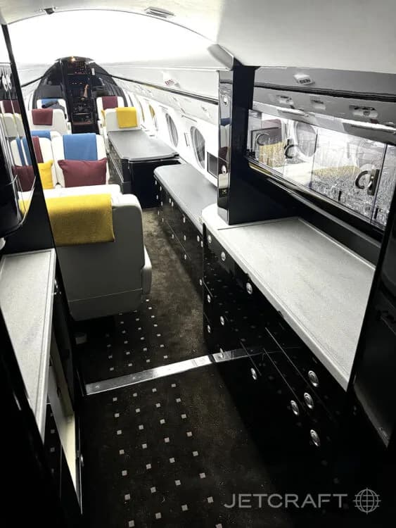 The interior of a private jet showing a galley or bar area with dark glossy cabinets, light countertops, and glassware.