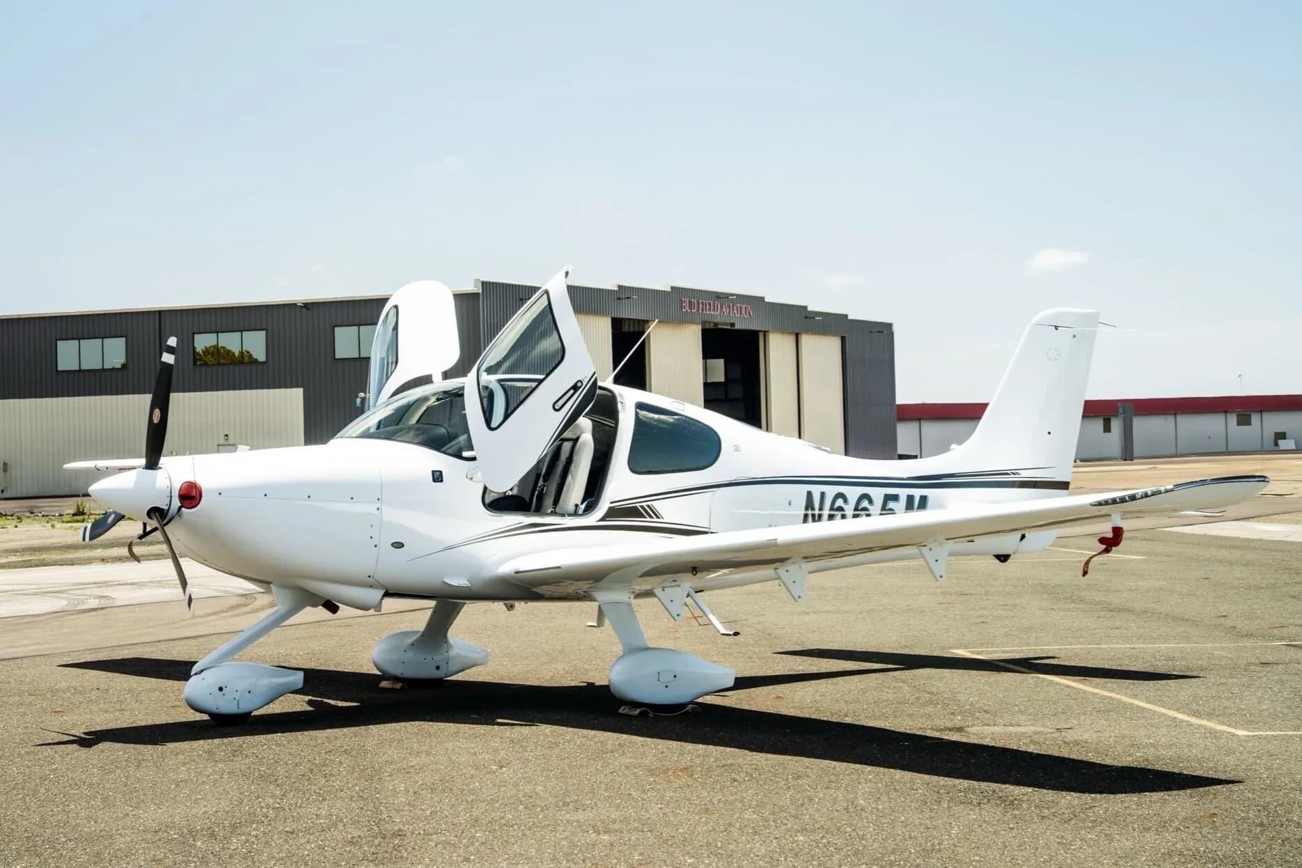 A white Cirrus SR22 aircraft with registration N665M parked on the tarmac with its doors open, with a hangar in the background.