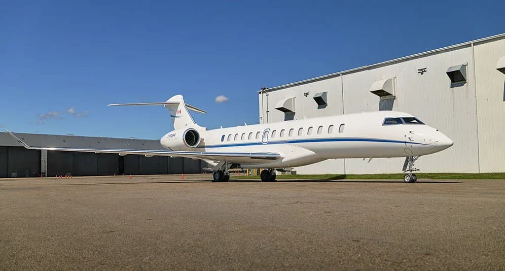 Side view of a white Bombardier Global business jet with blue stripes parked on the tarmac in front of a hangar.