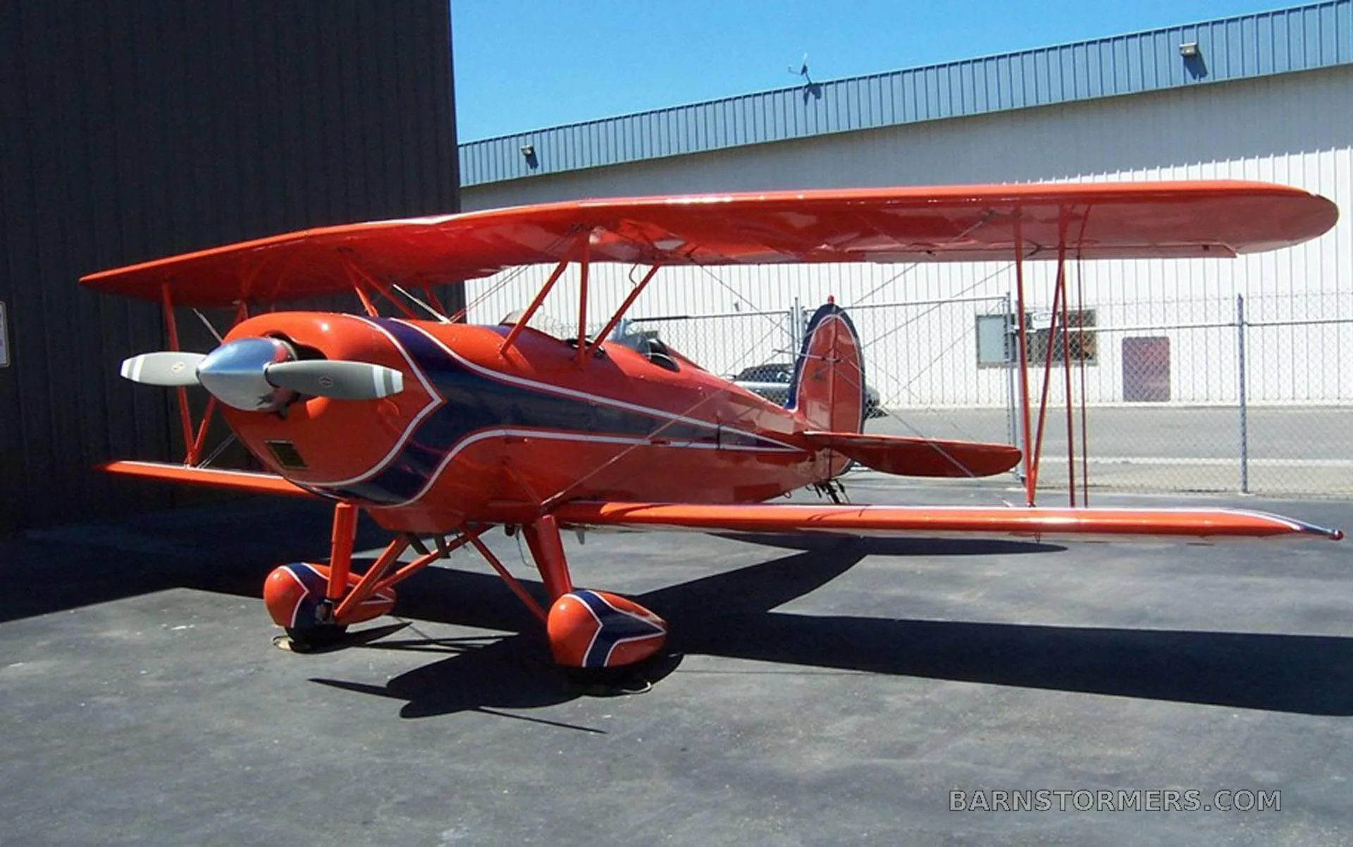 A red biplane with blue and white stripes is parked on an asphalt tarmac next to a building.