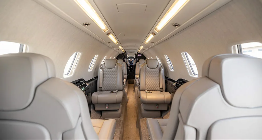 The interior cabin of a private jet with light gray quilted leather seats and overhead lighting.