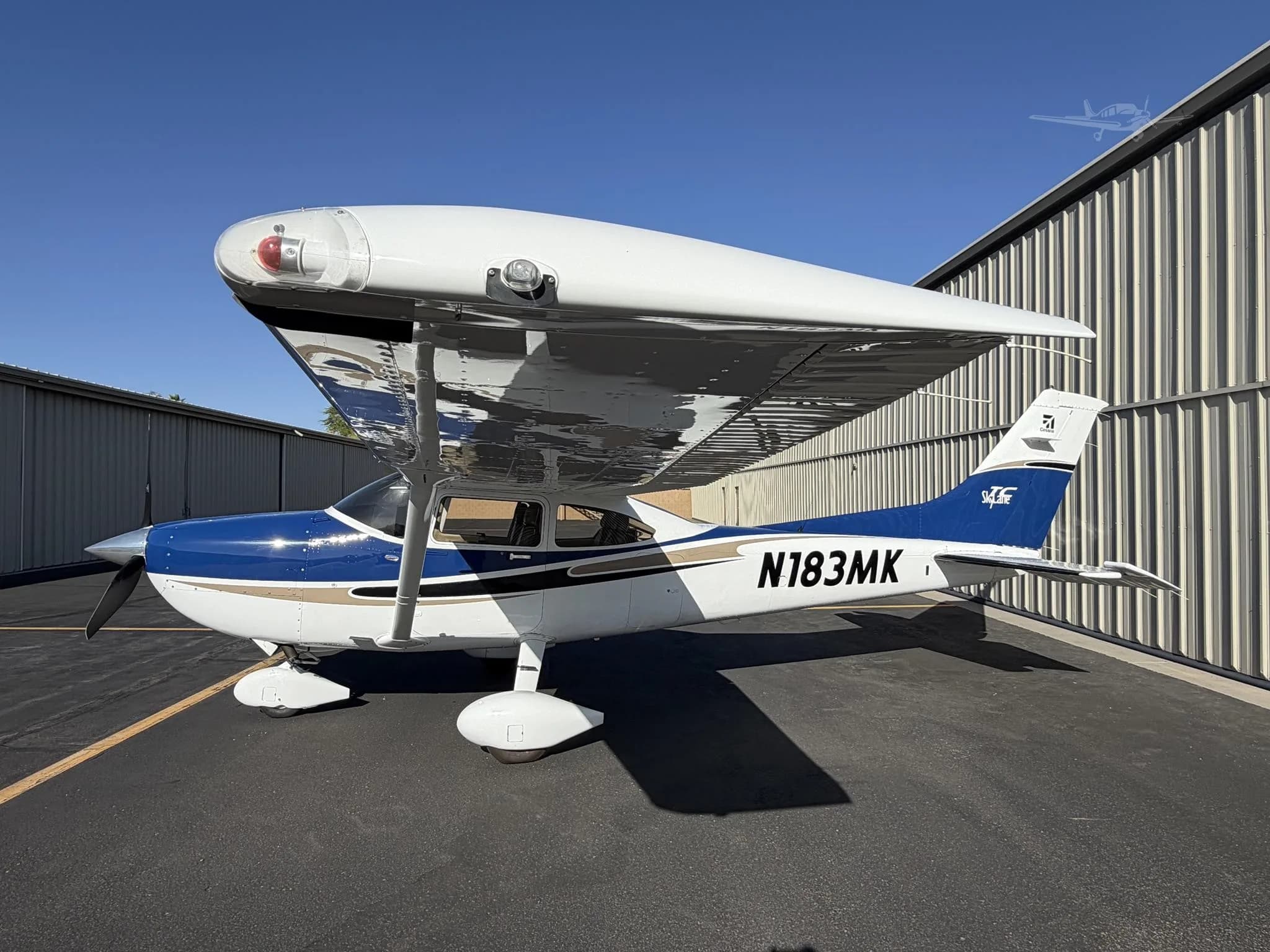 A blue and white Cessna 182 Skylane (N183MK) is parked on an asphalt ramp next to a hangar under a clear blue sky.