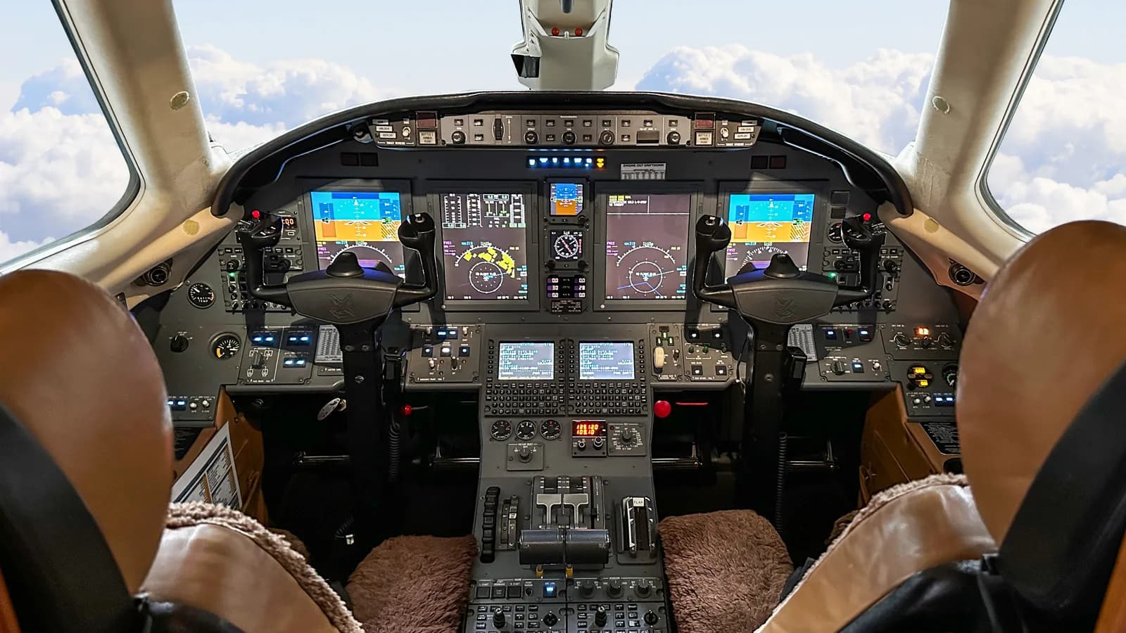 The cockpit of a private jet features multiple digital displays and controls, with clouds visible through the windshield.