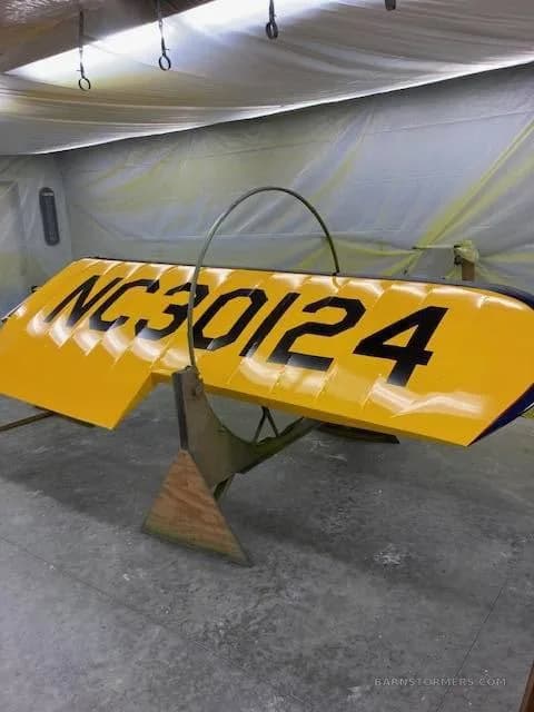 A yellow biplane wing with black registration NC 30124 is propped up in a workshop, possibly during painting or repair.