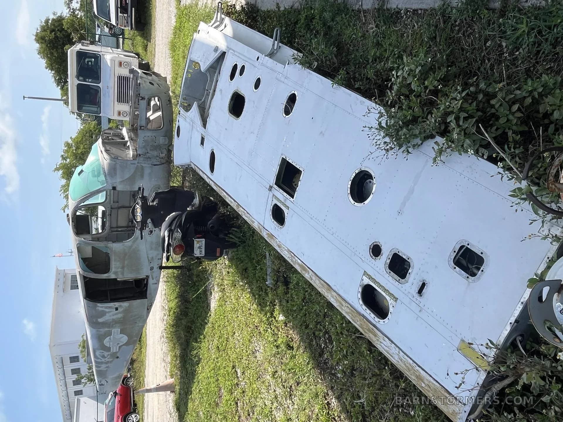 A disassembled aircraft fuselage and a detached wing section lie on the grass next to a dirt road.