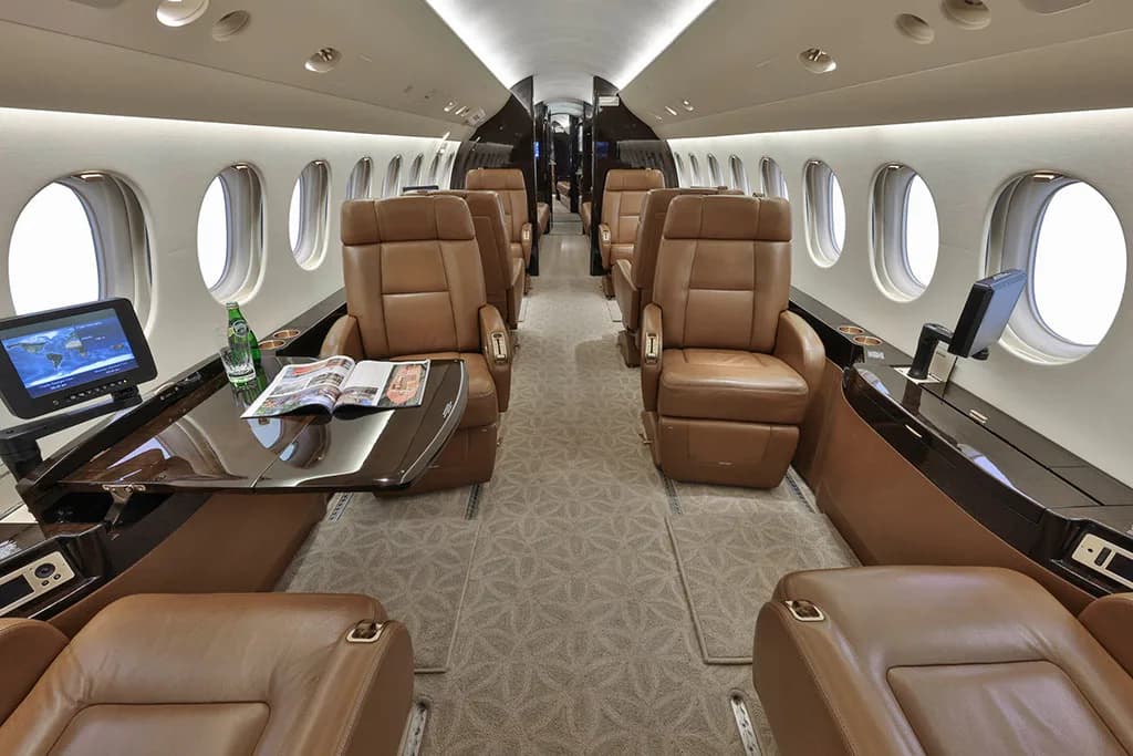 The interior cabin of a business jet shows tan leather seats, a table with a magazine and bottle, and large oval windows.
