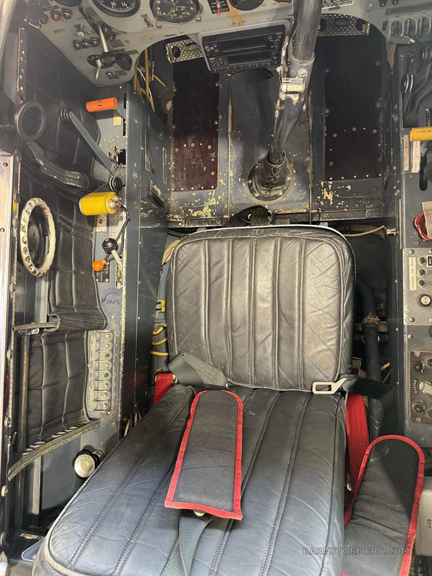 A close-up view of a black padded pilot's seat with red harness straps, surrounded by various controls and panels in an aircraft cockpit.