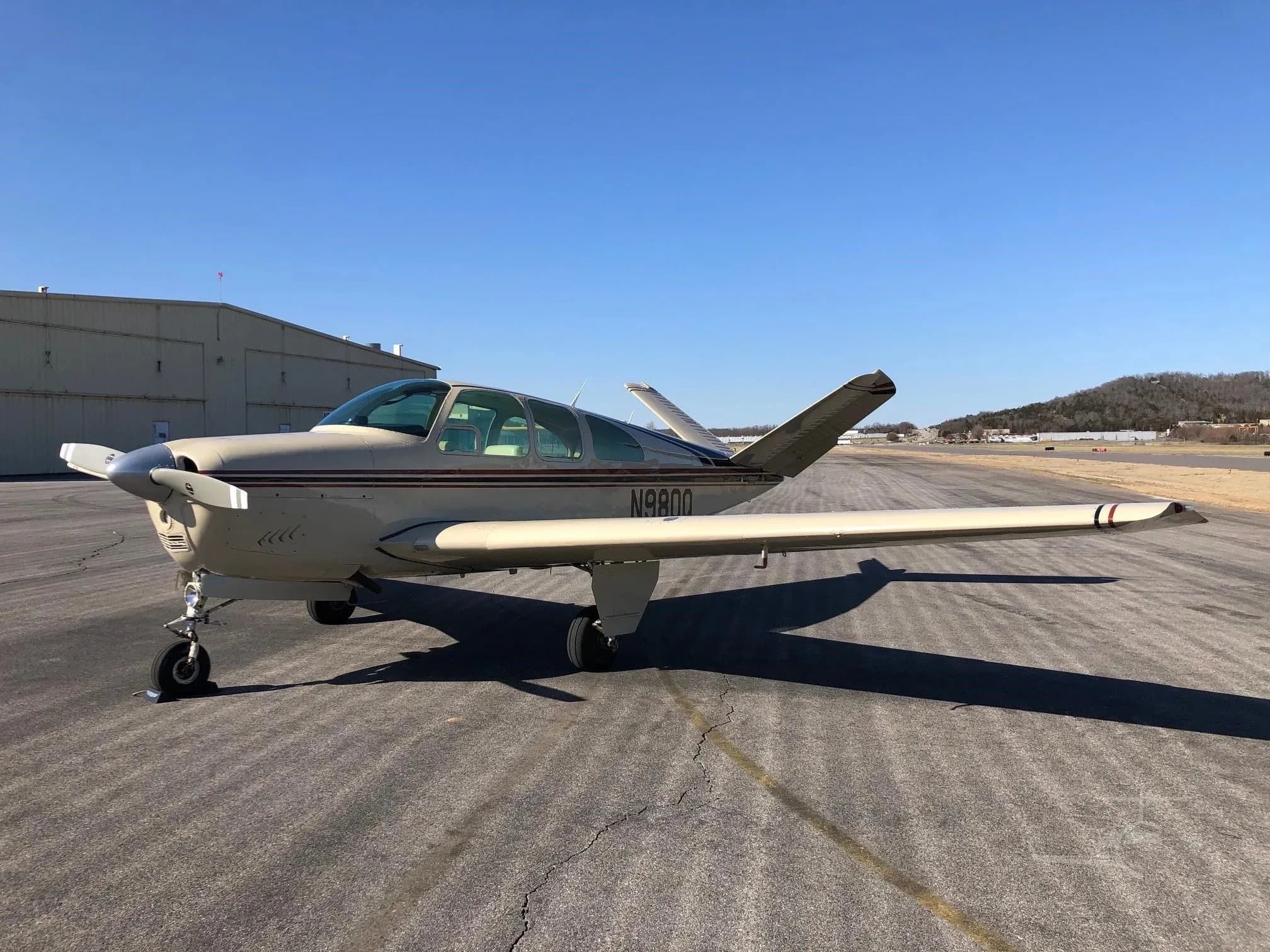 A front-quarter view of a beige Beechcraft Bonanza N980Q with red and black stripes parked on an asphalt runway near a hangar.