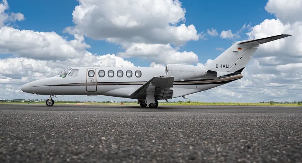 A white Cessna Citation business jet with black and brown stripes, registration D-IALI, parked on an asphalt runway under a blue sky with clouds.