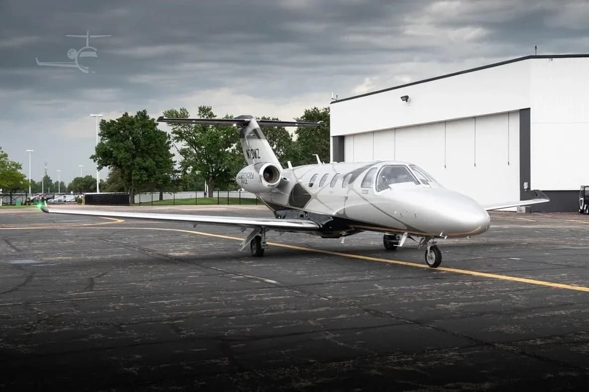 A wide shot of a silver and black Cessna Citation M2 jet (N70MZ) parked on an asphalt ramp with a hangar in the background.