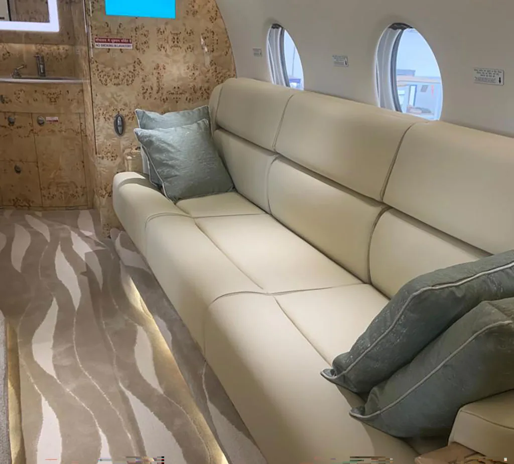 A long cream leather sofa with green pillows inside a private jet cabin, next to windows and patterned wood paneling.