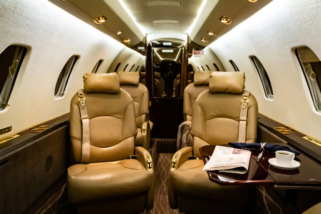 The luxurious interior cabin of a private jet features tan leather seats and a small table with a newspaper and coffee cup.
