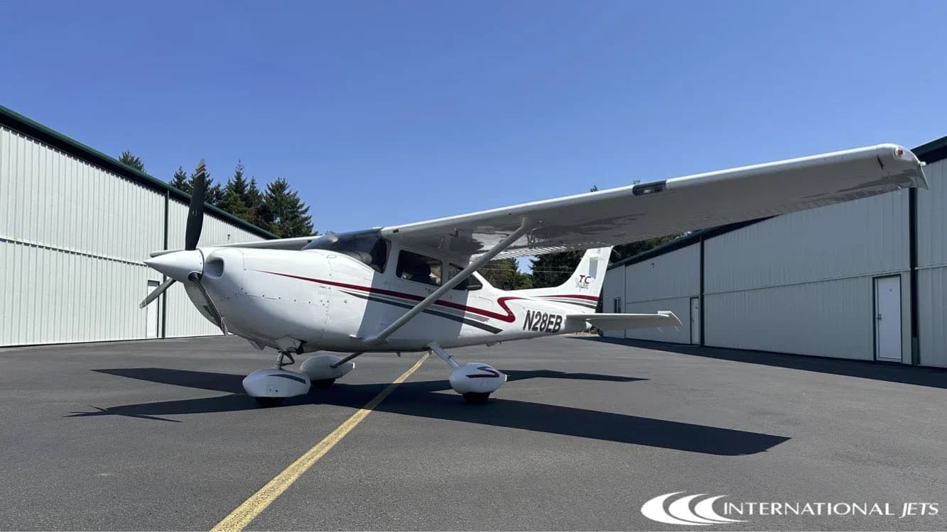 A white Cessna 182 Skylane with red and gray stripes, registration N28EB, parked on an asphalt ramp in front of a hangar.