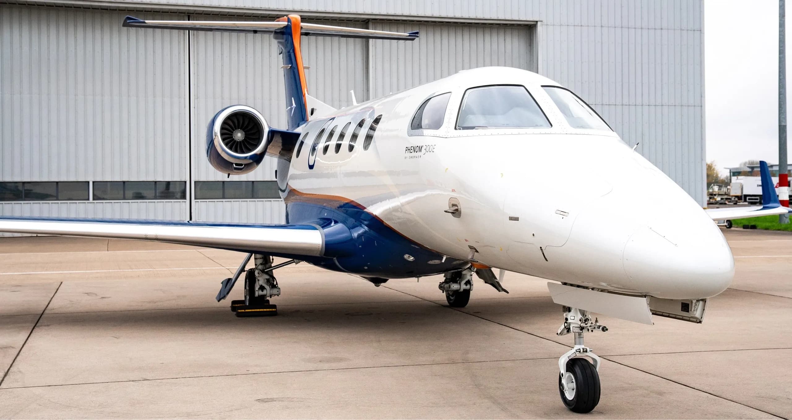 A close-up front-right view of a white and blue Embraer Phenom 300E business jet parked on a tarmac.