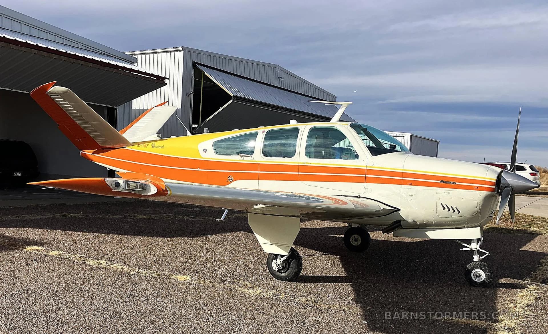 A front-quarter view of a white, orange, and yellow Beechcraft Bonanza N725AP parked on an airfield with an open hangar behind it.
