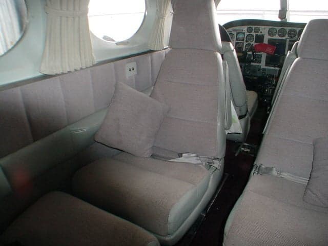 The aircraft cabin interior showing multiple rows of beige fabric seats and pleated window shades, with a partial view of the cockpit.