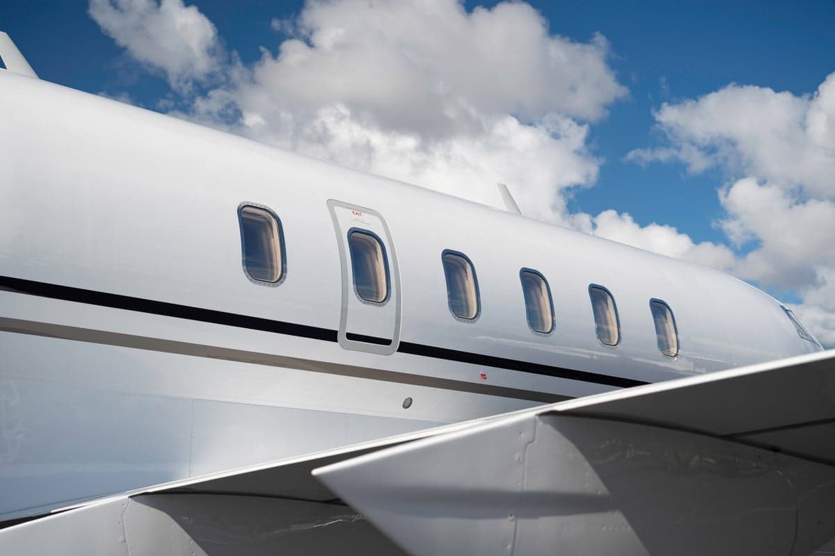 A side view of the white fuselage of a business jet showing passenger windows and an exit door, with a wing in the foreground.