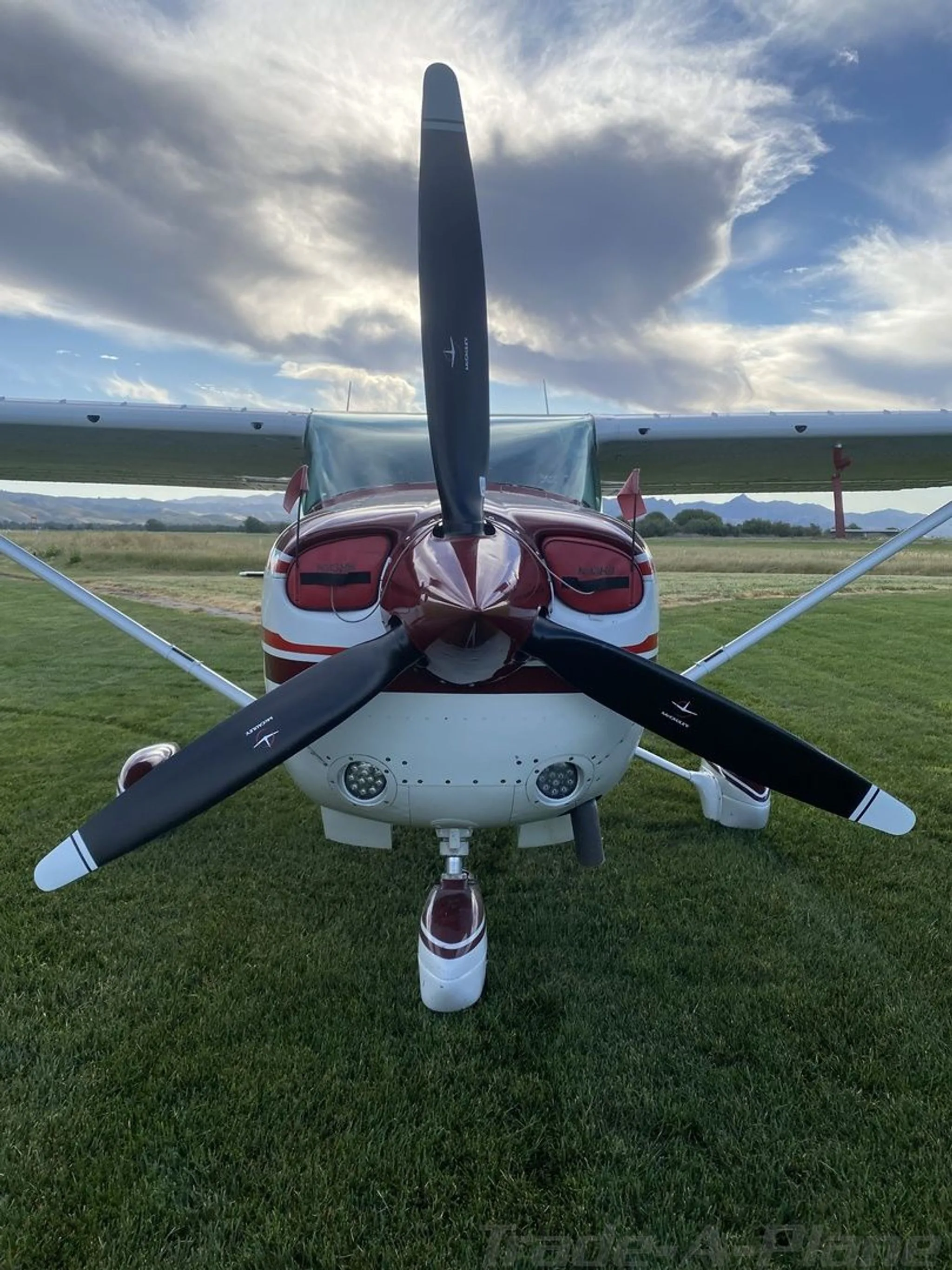 A close-up front view of the propeller and nose of a white Cessna 182 with red accents, parked on a grassy field.