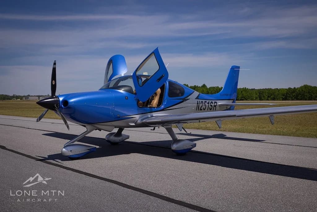 A blue and silver Cirrus SR20 aircraft (N251SR) with its doors open, parked on an airfield under a blue sky.