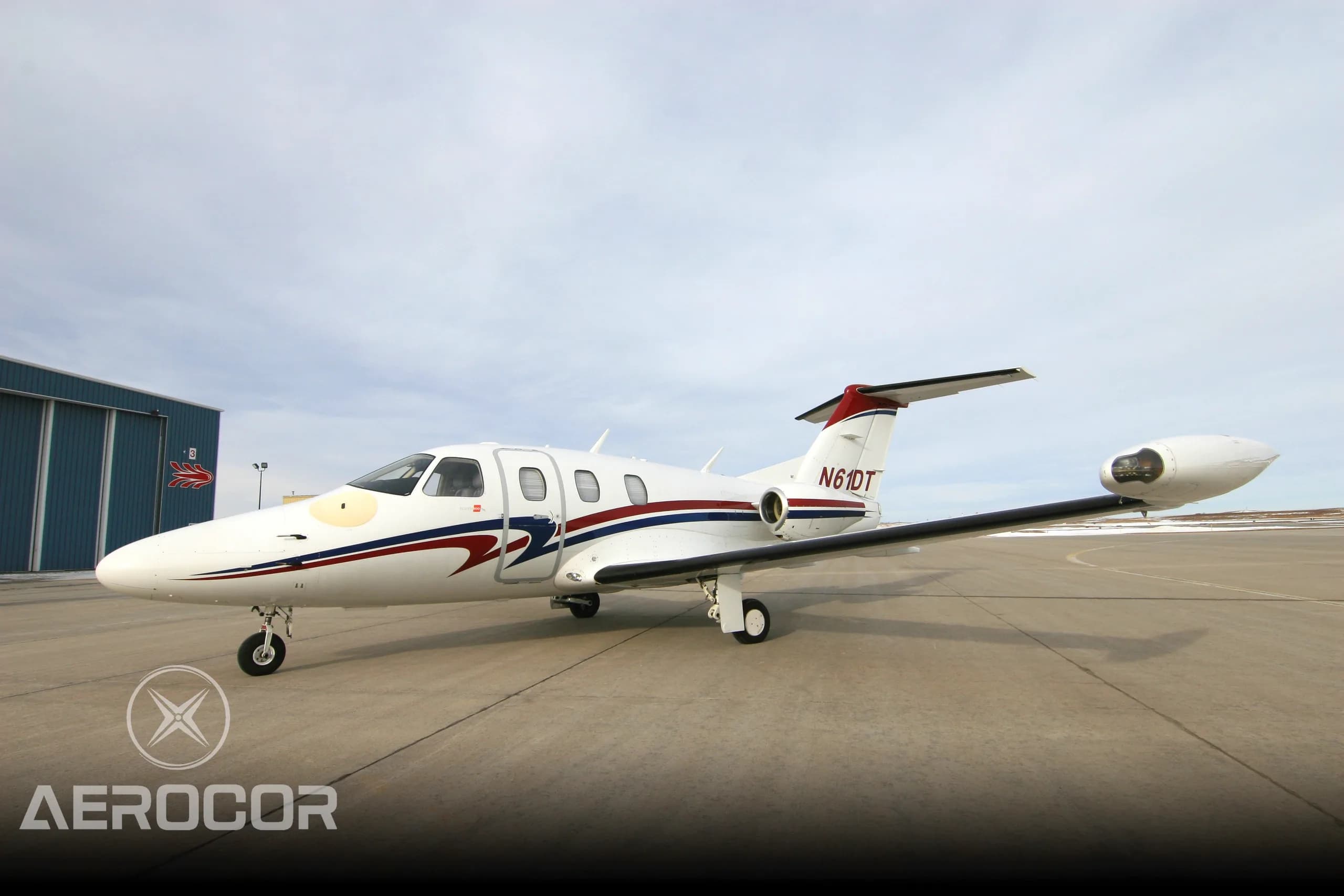 A white Eclipse 500 jet with red and blue stripes parked on an airport tarmac next to a hangar.