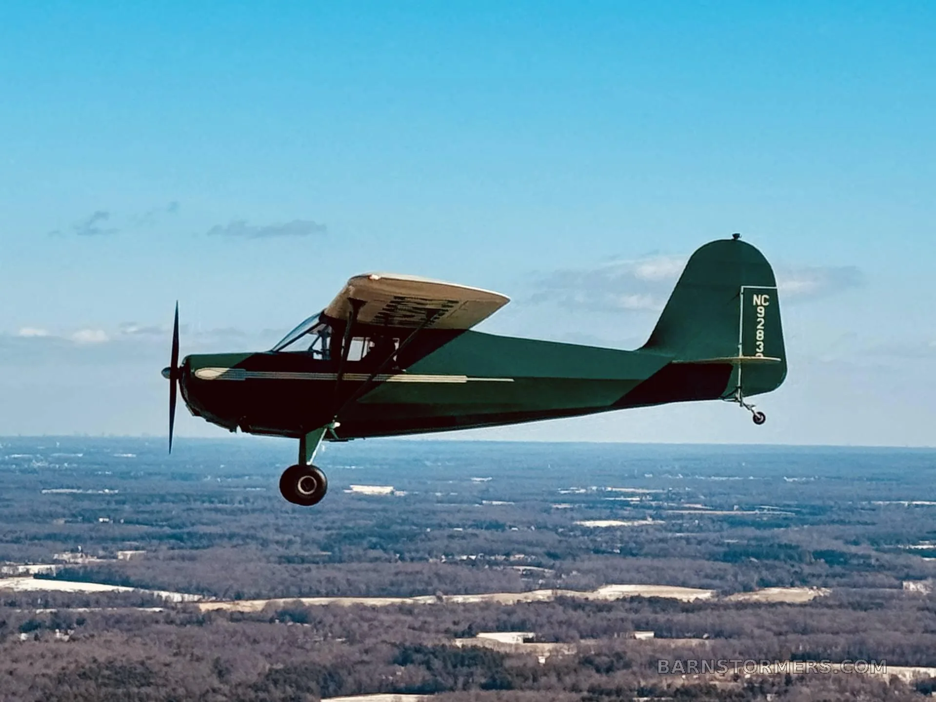 A green and cream vintage aircraft with tail number NC92833 is flying over a wooded landscape.